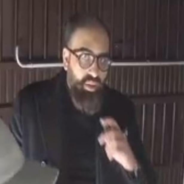 Mann mit Bart und Brille in dunkler Kleidung steht vor einer Holzwand.