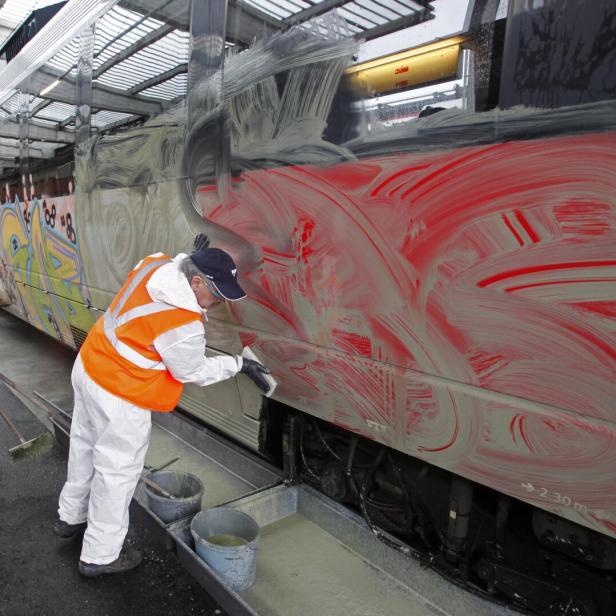 Zwei Personen in Schutzkleidung und Warnwesten reinigen Graffiti von einem Zug mit Bürsten und Reinigungsmitteln.
