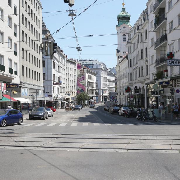 Mehrspurige Straße mit Autos, Straßenbahnschienen, Geschäften, Menschen auf Gehwegen und einer Kirche mit grünem Turm im Hintergrund.