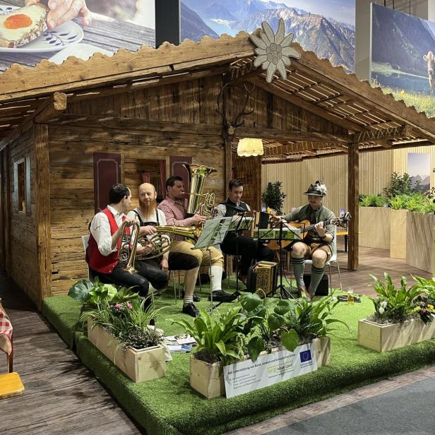 Vier Musiker in traditioneller Tracht spielen Blasinstrumente und Gitarre vor einer Holzhütte mit Alpenkulisse.