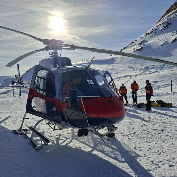 Ein Rettungshubschrauber steht im verschneiten Gebirge, daneben mehrere Personen in Ausrüstung.