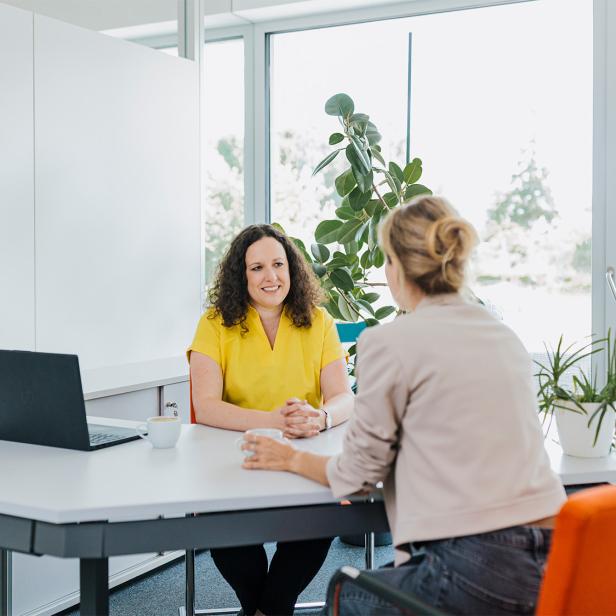 Zwei Frauen sitzen sich an einem Schreibtisch in einem hellen Büro gegenüber und unterhalten sich.