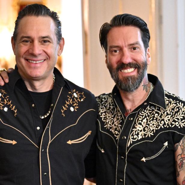 Zwei Männer mit bestickten schwarzen Hemden stehen lächelnd nebeneinander, einer hat sichtbare Tattoos am Arm.