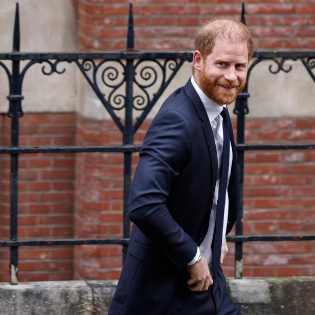 Prinz Harry in Anzug und Krawatte lächelt vor einem Backsteingebäude.
