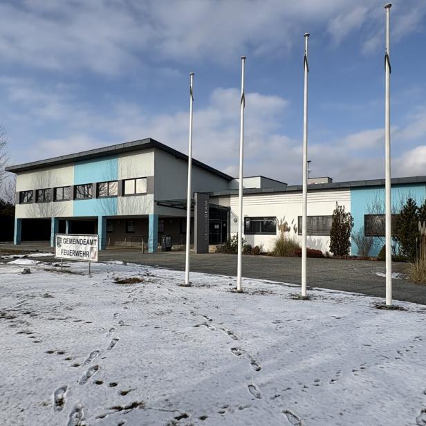 Ein Gebäude des Gemeindeamts/Feuerwehr mit vier Fahnenmasten steht vor einem teilweise schneebedeckten Feld.
