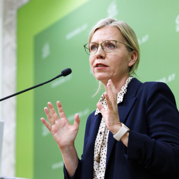 Gewessler gestikuliert vor einem grünen Hintergrund mit Mikrofon.