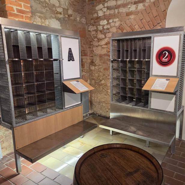 Zwei Metallregale mit nummerierten Fächern und Infotafeln stehen vor einer Backsteinwand in einem historischen Raum.