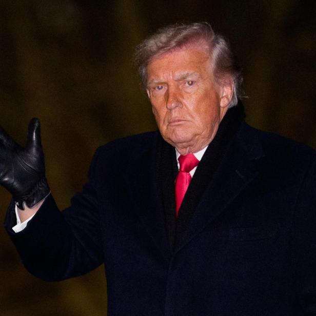 Donald Trump in dunklem Mantel und roter Krawatte grüßt mit einem schwarzen Handschuh.