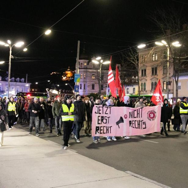 Demonstrierende mit Transparenten und roten Fahnen ziehen nachts durch eine Grazer Straße.