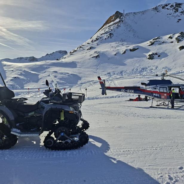 Ein Quad mit Ketten und ein Polizei-Hubschrauber vom Typ OE-BXN stehen vor einer verschneiten Berglandschaft.