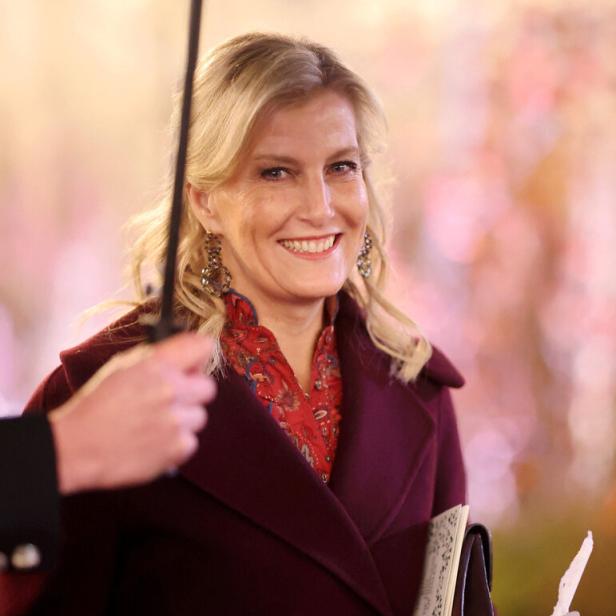 Eine lächelnde Herzogin Sophie mit blonden Haaren trägt einen burgunderroten Mantel und hält eine Kerze und eine Broschüre.