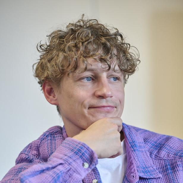 Tim Bendzko mit lockigen blonden Haaren trägt ein lila Hemd und blickt nachdenklich zur Seite.
