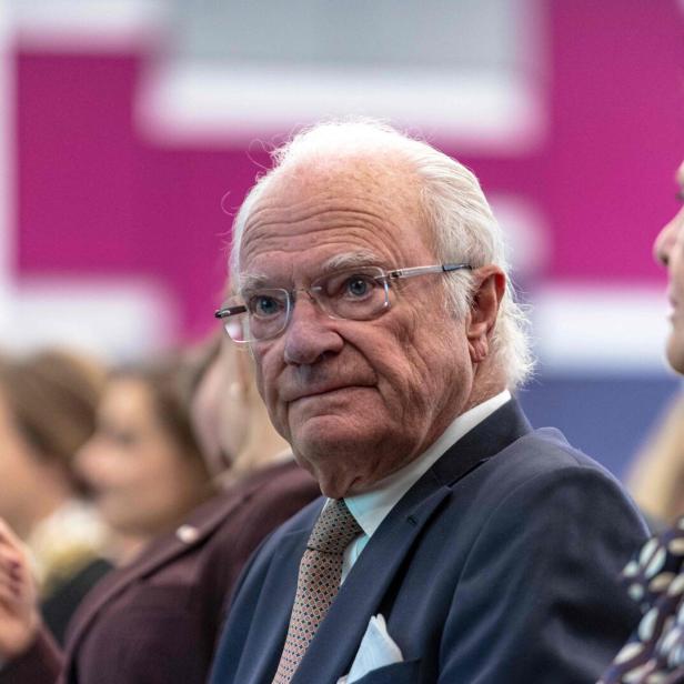 König Carl XVI. Gustaf mit Brille und Anzug sitzt neben Königin Silvia bei einer Veranstaltung.