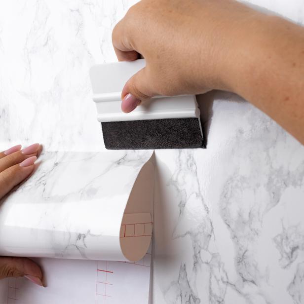 Hände bringen eine Tapete in Marmoroptik mit einer Rakel an einer Wand an.