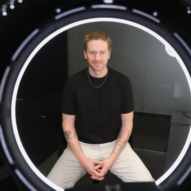 Ein Mann mit Tattoos sitzt vor einer leuchtenden Ringlampe und schaut direkt nach vorne.