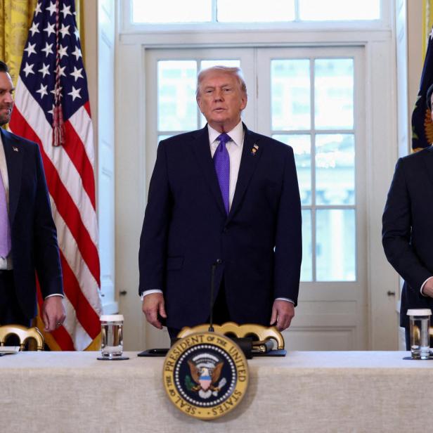 Donald Trump steht mit zwei Männern, JD Vance und Marco Rubio, vor US-Flaggen.