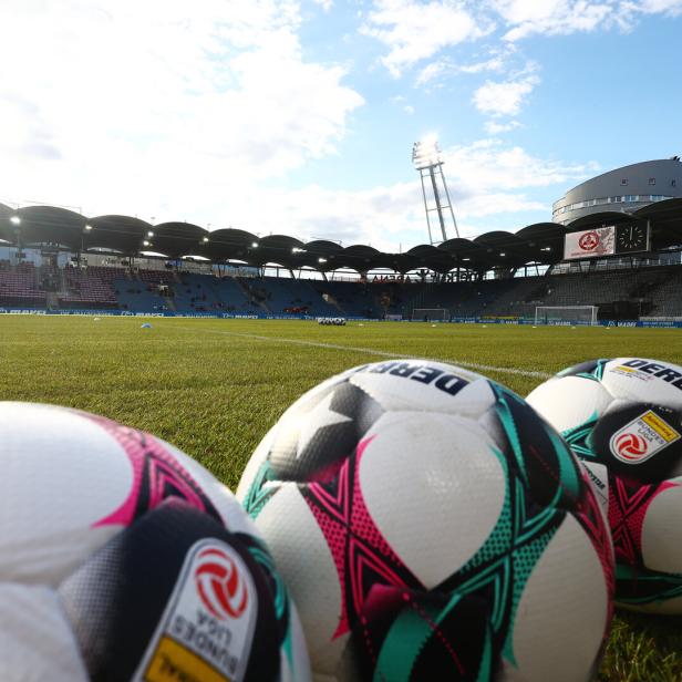 Mehrere Fußbälle liegen am Spielfeldrand eines leeren Fußballstadions bei Tageslicht.