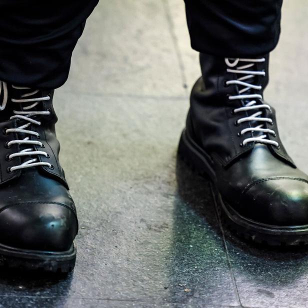 Zwei schwarze, geschnürte Stiefel auf einem dunklen Boden, getragen von einer Person mit schwarzer Hose.