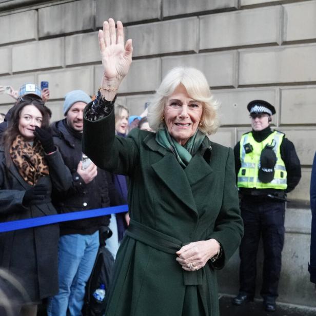 Camilla, in einem grünen Mantel, winkt einer Menschenmenge vor einem Gebäude zu.