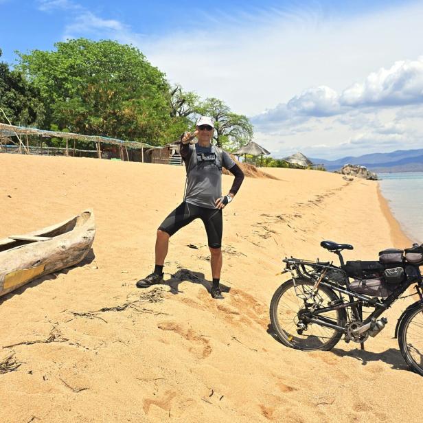 Eine Person steht auf einem Sandstrand neben einem Fahrrad und einem Einbaum, im Hintergrund Bäume und Wasser.