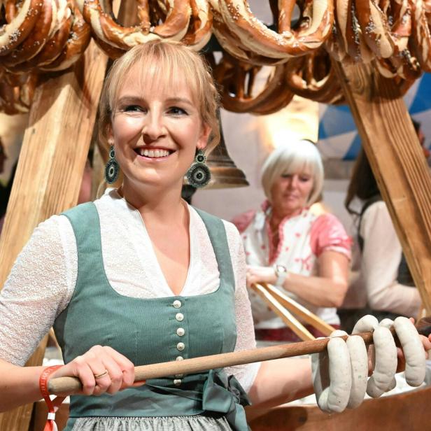Eine Frau in Tracht hält eine große Holzstange mit Weißwürsten, im Hintergrund hängen viele Brezeln.
