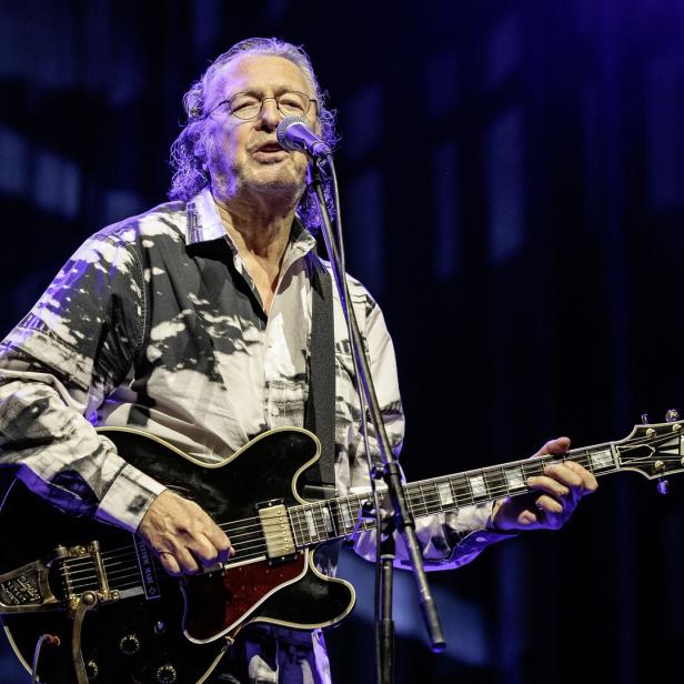 Ein älterer Mann - Peter Cornelius - mit Brille spielt E-Gitarre und singt auf einer Bühne vor dunklem Hintergrund.