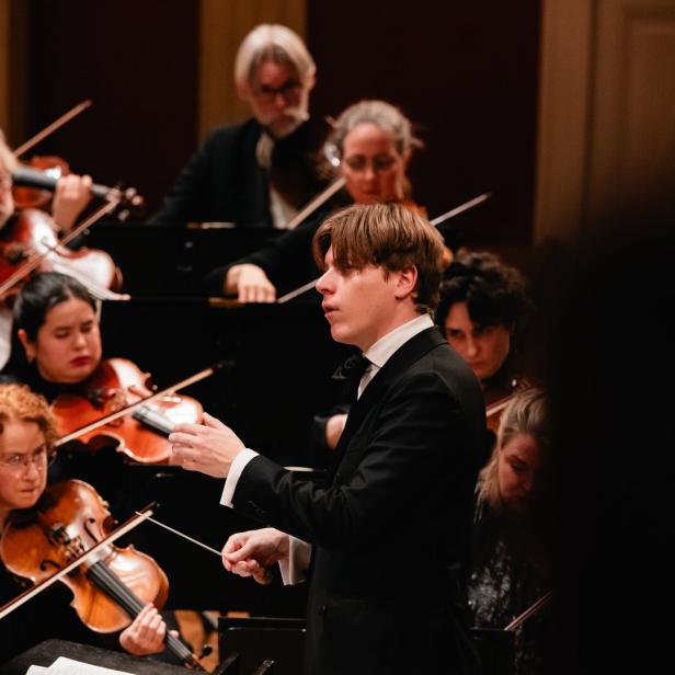 Ein Dirigent - Klaus Mäkelä -  leitet ein Orchester, dessen Musiker konzentriert Streichinstrumente spielen.