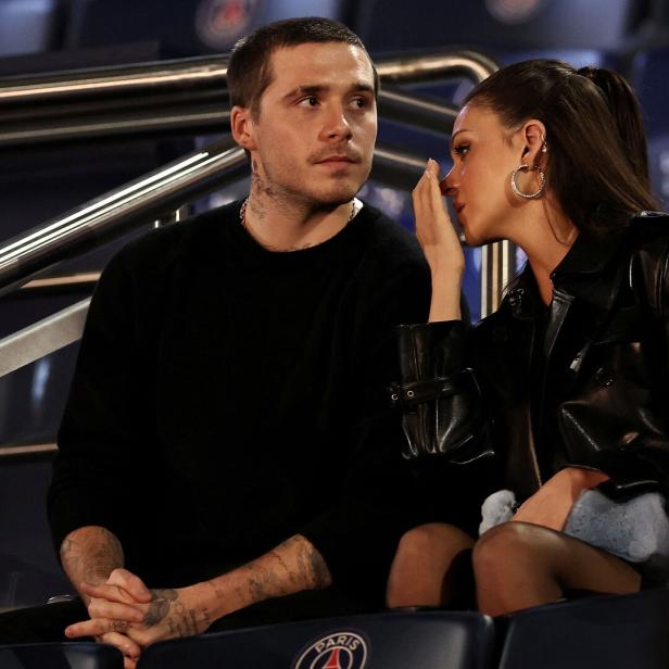 Brooklyn Beckham und Nicola Peltz sitzen nebeneinander. Sie flüstert ihm etwas ins Ohr, während er aufmerksam zuhört.