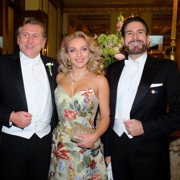 Drei elegant gekleidete Personen posieren lächelnd nebeneinander, zwei Männer im Frack und eine Frau im floralen Abendkleid.