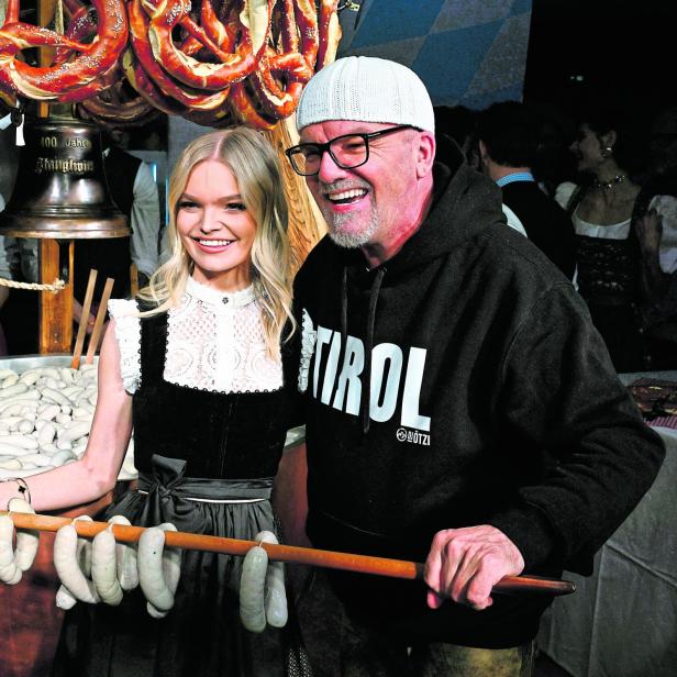 Eine Frau in Dirndl und ein Mann mit Tirol-Pullover halten gemeinsam einen Holzstab mit Weißwürsten, im Hintergrund Brezeln.