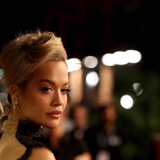 Eine elegant gekleidete Frau mit hochgesteckten blonden Haaren blickt über die Schulter, im Hintergrund unscharfe Lichter.