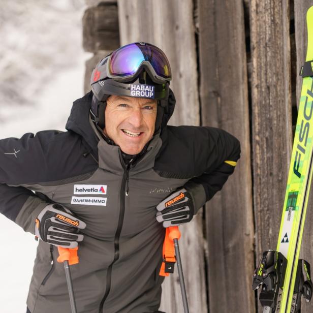 Ein Skifahrer in Winterkleidung lehnt mit Skistöcken an einer Holzwand, neben ihm stehen gelbe Skier.