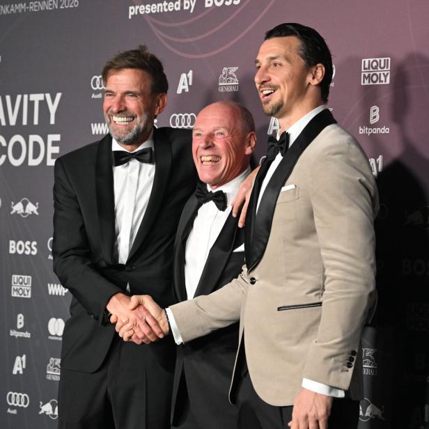 Drei Männer in festlicher Kleidung lachen und posieren gemeinsam vor einer Sponsorenwand.