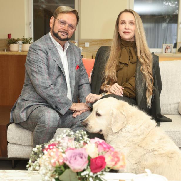 Ein Mann und eine Frau sitzen auf einem Sofa, vor ihnen ein großer, heller Hund und ein Blumenstrauß auf dem Tisch.