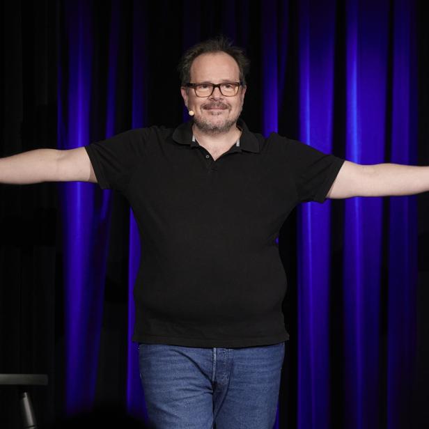 Ein Mann mit Brille und Mikrofon steht mit ausgebreiteten Armen auf einer Bühne vor dunklem Vorhang, daneben ein Hocker mit Wasserflasche.