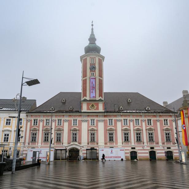 Ein großes historisches Gebäude mit Uhrturm, rosa Fassade und mehreren Flaggen davor auf einem leeren Platz.