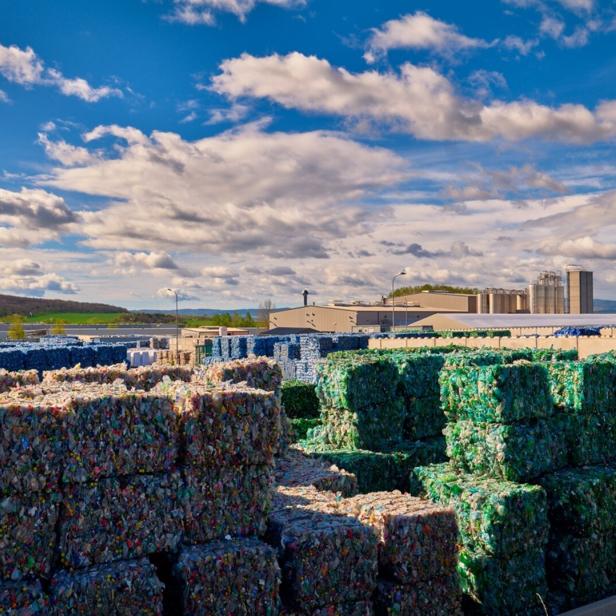 Gestapelte, gepresste Ballen aus recyceltem Kunststoff lagern im Freien vor einer Industrieanlage.