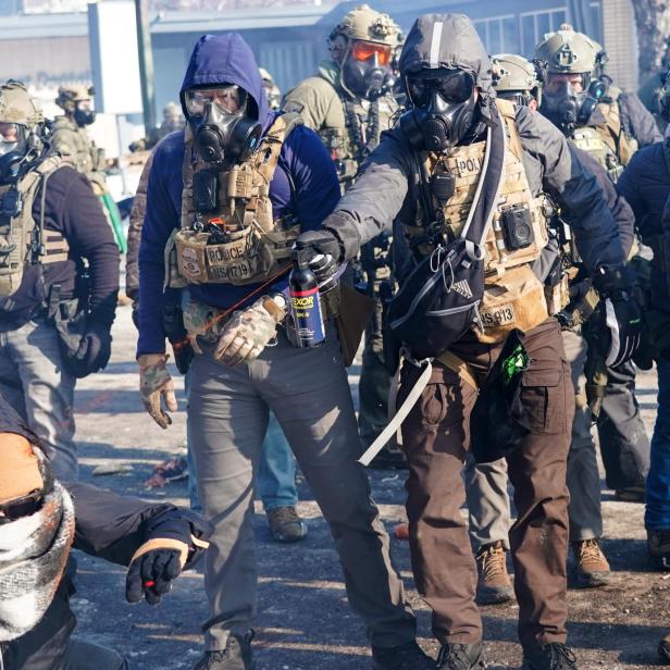 Eine Gruppe von Polizisten in Schutzausrüstung steht auf einer Straße, während eine Person am Boden kauert.