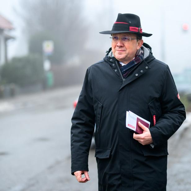 Ein Mann mit Hut und Jacke hält Papiere in der Hand und geht auf einer nebligen Straße.
