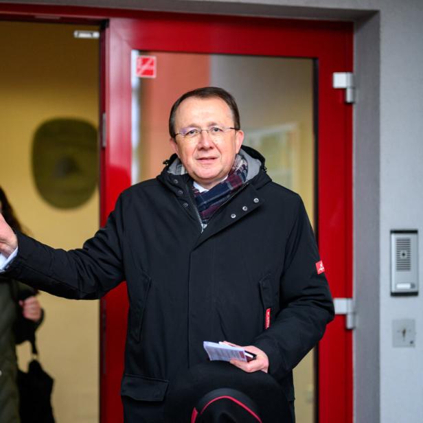Ein Mann mit Brille und Jacke steht vor einer roten Tür mit Hinweisen zum Wahllokal.