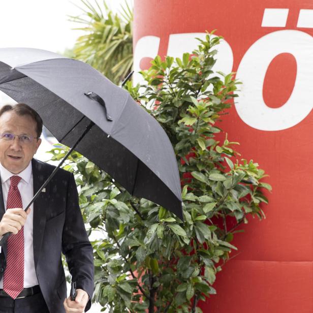 Ein Mann mit Anzug und Krawatte hält einen Regenschirm im Regen vor einem roten Hintergrund mit einem weißen "Ö".