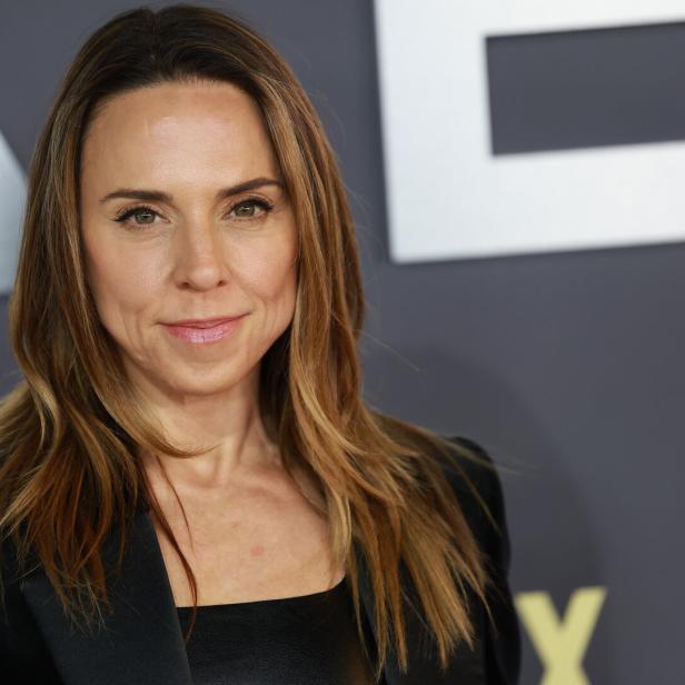 Melanie Chisholm lächelt mit offenem Haar und schwarzem Blazer vor einem grauen Hintergrund.