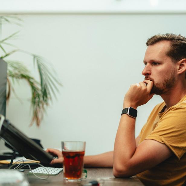 Ein Mann sitzt nachdenklich an einem Schreibtisch vor zwei Monitoren, mit einem Glas Getränk daneben.