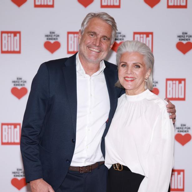 Birgit Schrowange und Frank Spothelfer stehen lächelnd Arm in Arm vor einer Wand mit roten Herzen und Logos.