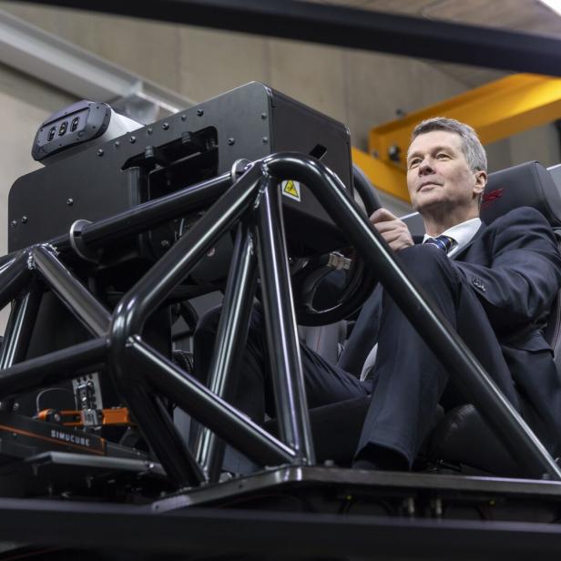 Ein Mann im Anzug sitzt in einem Fahrsimulator mit schwarzem Stahlrahmen und blickt nach oben.