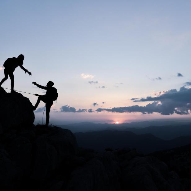 Zwei Wanderer helfen sich beim Aufstieg auf einen Felsen im Sonnenuntergang.