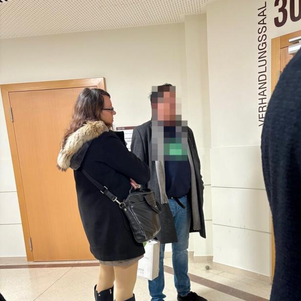 Zwei Personen stehen in einem Flur vor einer Tür mit der Aufschrift „Verhandlungssaal“.