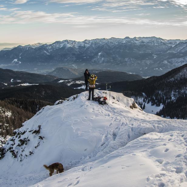 Zwei Wanderer mit Hund auf verschneitem Berggipfel, im Hintergrund das Karwendelgebirge.