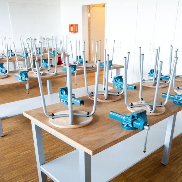 Mehrere Werkbänke mit Schraubstöcken, auf denen umgedrehte Hocker stehen, in einem hellen Raum mit Holzfußboden.