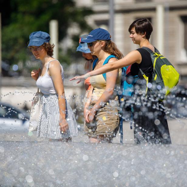 Drei junge Frauen stehen an einem sonnigen Tag an einem sprudelnden Brunnen und schauen auf das Wasser.